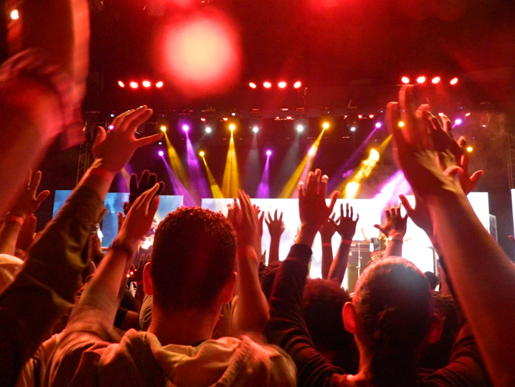 A vibrant crowd illuminated by lavender and light blue stage lights, hands raised in celebration.