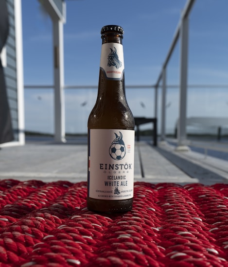 A bottle of Einstök Icelandic White Ale is placed on a textured red surface. The setting appears to be outdoors, with a clear blue sky and blurred elements suggesting a patio or deck area with glass railings.