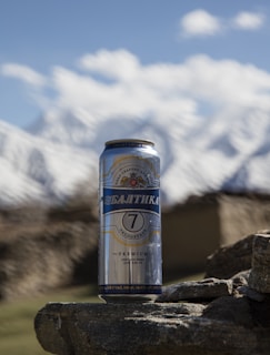 A metallic can of Baltika 7 beer is placed on a rough stone surface. In the background, snow-capped mountains and a clear blue sky are visible, creating a natural and refreshing setting.