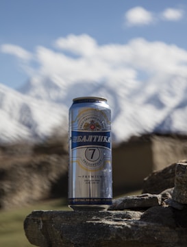 A metallic can of Baltika 7 beer is placed on a rough stone surface. In the background, snow-capped mountains and a clear blue sky are visible, creating a natural and refreshing setting.