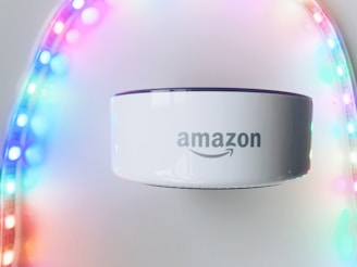 A white Amazon device, possibly a smart speaker, is positioned in the center with a soft focus on colorful LED strip lights forming an arch around it. The LED lights display a gradient of colors including purple, pink, blue, and green.
