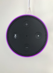 A circular electronic device with a dark surface featuring control buttons, including volume and microphone icons. It has a glowing pinkish-purple LED ring around the edge and is connected via a white cable.