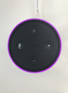 A circular electronic device with a dark surface featuring control buttons, including volume and microphone icons. It has a glowing pinkish-purple LED ring around the edge and is connected via a white cable.
