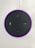A circular electronic device with a dark surface featuring control buttons, including volume and microphone icons. It has a glowing pinkish-purple LED ring around the edge and is connected via a white cable.