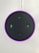 A circular electronic device with a dark surface featuring control buttons, including volume and microphone icons. It has a glowing pinkish-purple LED ring around the edge and is connected via a white cable.