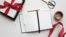 A flat lay arrangement of a gift box wrapped with a red ribbon next to an open planner or calendar. Scissors with a gold handle, a wooden bowl containing paper clips, and a golden pen are also present. A long red ribbon and small star-shaped decorations are scattered around the scene.