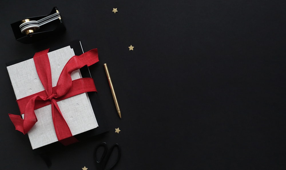 white gift box with red ribbon beside pencil, scissors, and tape dispenser