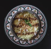 A decorative plate features a traditional Middle Eastern dessert garnished with a sprinkle of crushed nuts and a mint leaf, surrounded by a design of intricate patterns with vibrant colors.