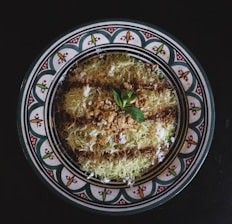 A decorative plate features a traditional Middle Eastern dessert garnished with a sprinkle of crushed nuts and a mint leaf, surrounded by a design of intricate patterns with vibrant colors.