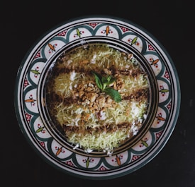 A decorative plate features a traditional Middle Eastern dessert garnished with a sprinkle of crushed nuts and a mint leaf, surrounded by a design of intricate patterns with vibrant colors.