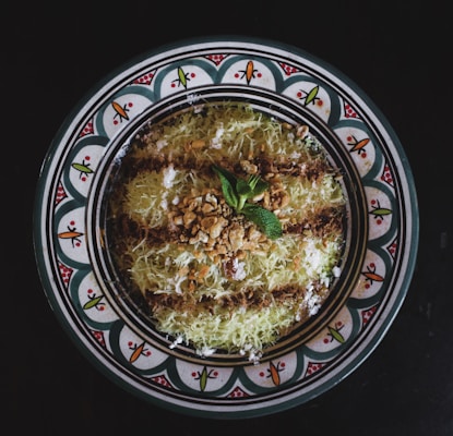 A decorative plate features a traditional Middle Eastern dessert garnished with a sprinkle of crushed nuts and a mint leaf, surrounded by a design of intricate patterns with vibrant colors.
