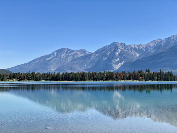 A serene landscape painting of a mountain lake surrounded by pine trees under a clear blue sky.