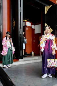 Two individuals are wearing traditional Japanese kimonos, standing by the entrance of a building with red and black detailing. One person is holding a smartphone, and the other appears to be in conversation using a phone. The atmosphere seems casual and contemporary within a traditional setting.