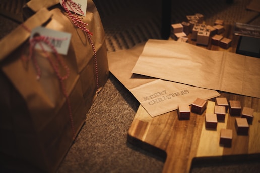 A festive scene with two brown paper gift bags, each adorned with red and white twine and tags. Wooden stamps and a paper with the words 'Merry Christmas' are placed on a wooden surface. Various small, cube-shaped wooden blocks are scattered around.