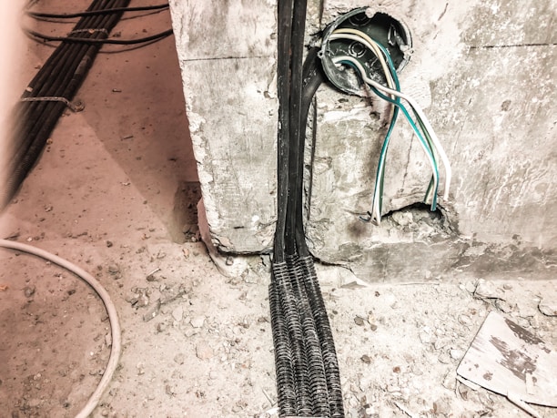 A construction site is depicted with exposed electrical wires protruding from a circular conduit in a rough concrete wall. The floor is covered in dust and debris, and there are multiple cables organized in a black mesh covering running along the floor.