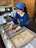 Photo of a woman in a traditional apron arranging sweets in her shop with milk bottles on the counter.