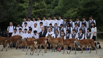 A group of people, likely students and two older adults, are dressed in matching uniforms and positioned in several rows for a group photo. They are outdoors, surrounded by greenery and trees, with several deer standing in front of them. The people seem to be posing formally, and the setting appears serene and natural.