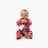 A baby wearing festive red pajamas decorated with holiday patterns sits on a white background. The baby is holding a black card or object with both hands and seems to be chewing on it. There is a grey cord attached to the object and a white tag hanging from it.