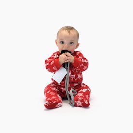 A baby wearing festive red pajamas decorated with holiday patterns sits on a white background. The baby is holding a black card or object with both hands and seems to be chewing on it. There is a grey cord attached to the object and a white tag hanging from it.