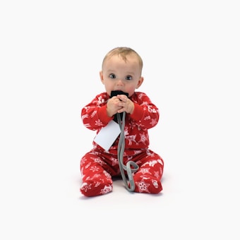 A baby wearing festive red pajamas decorated with holiday patterns sits on a white background. The baby is holding a black card or object with both hands and seems to be chewing on it. There is a grey cord attached to the object and a white tag hanging from it.