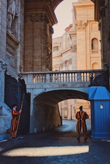 Two guards in vibrant, traditional uniforms stand at either side of an ornate archway in an architectural setting. The scene includes a blend of shadow and light, highlighting the grandeur of stone columns and intricate carvings.