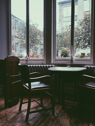 A cozy interior with a round wooden table and two chairs positioned near tall windows. The view through the windows shows an urban street with buildings and trees. The lighting is soft and gives the space a warm, inviting atmosphere.