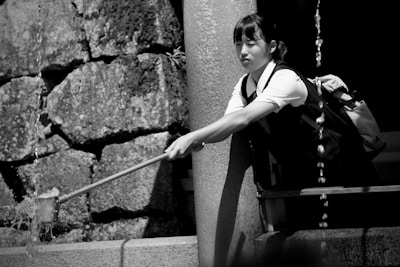 A person in a school uniform reaches out with a ladle to catch water from a stream in a traditional setting with stone and concrete elements.