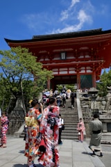 A group of people are walking up the stone steps leading to a traditional Japanese temple gate, surrounded by lush greenery. Several women are wearing vibrant kimonos with intricate patterns, and the clear blue sky provides a striking backdrop.
