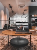 A modern, spacious interior with a cozy, stylish atmosphere. A round wooden table in the foreground holds an open laptop displaying a scenic landscape. The room has a large window in the background allowing natural light, accompanied by contemporary furniture and decor elements.