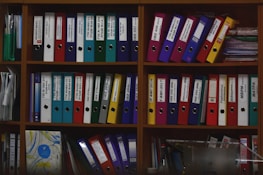 A wooden shelf is filled with various colorful ring binders and folders neatly organized. The binders have labels with text on them and are stacked both horizontally and vertically. Some loose papers and additional documents are tucked among the folders. The scene suggests an office or study environment with a focus on organization.