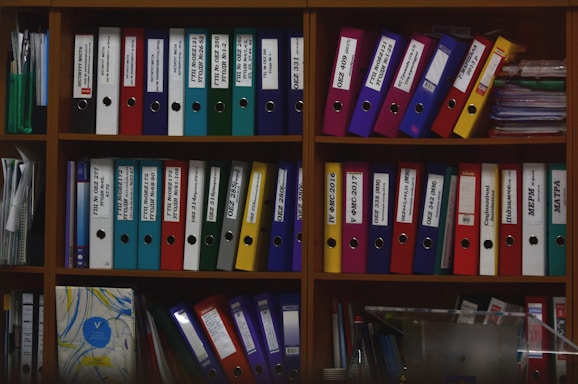 A wooden shelf is filled with various colorful ring binders and folders neatly organized. The binders have labels with text on them and are stacked both horizontally and vertically. Some loose papers and additional documents are tucked among the folders. The scene suggests an office or study environment with a focus on organization.