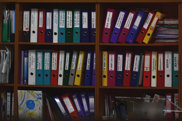 A wooden shelf is filled with various colorful ring binders and folders neatly organized. The binders have labels with text on them and are stacked both horizontally and vertically. Some loose papers and additional documents are tucked among the folders. The scene suggests an office or study environment with a focus on organization.