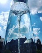 A large glass structure containing swirling water, resembling a vortex, stands against a bright blue sky with scattered clouds. Reflections of surrounding buildings and a clock tower are visible on the glass surface, creating an interplay of light and reflections.