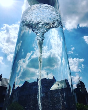 A large glass structure containing swirling water, resembling a vortex, stands against a bright blue sky with scattered clouds. Reflections of surrounding buildings and a clock tower are visible on the glass surface, creating an interplay of light and reflections.