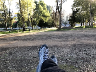 A person is relaxing outdoors with their legs stretched out, wearing sneakers. The view includes a dirt path and trees nearby, with buildings visible in the background. Sunlight filters through the foliage, creating a serene atmosphere.