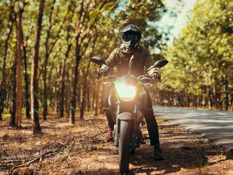A person wearing a helmet and protective gear is sitting on a motorcycle in a sunlit forest area. The motorcycle's headlight is turned on, and the person is facing directly forward, surrounded by tall trees with green leaves. The scene conveys a sense of adventure and exploration on a winding road.