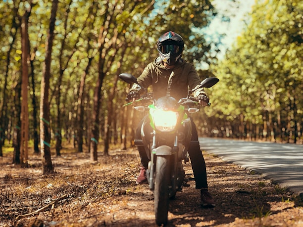 A person wearing a helmet and protective gear is sitting on a motorcycle in a sunlit forest area. The motorcycle's headlight is turned on, and the person is facing directly forward, surrounded by tall trees with green leaves. The scene conveys a sense of adventure and exploration on a winding road.