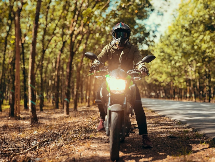 A person wearing a helmet and protective gear is sitting on a motorcycle in a sunlit forest area. The motorcycle's headlight is turned on, and the person is facing directly forward, surrounded by tall trees with green leaves. The scene conveys a sense of adventure and exploration on a winding road.