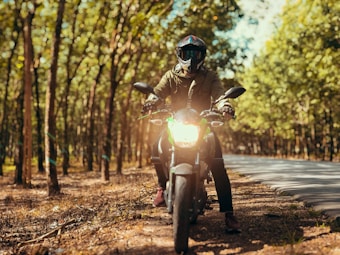 A person wearing a helmet and protective gear is sitting on a motorcycle in a sunlit forest area. The motorcycle's headlight is turned on, and the person is facing directly forward, surrounded by tall trees with green leaves. The scene conveys a sense of adventure and exploration on a winding road.