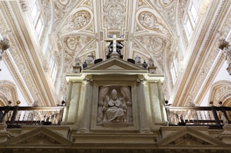 An ornate interior view featuring intricately decorated ceilings with elaborate carvings and moldings. The centerpiece is a sculptural relief of a seated figure within a niche, surrounded by detailed architectural elements. Natural light streams through windows, enhancing the grandeur of the setting.