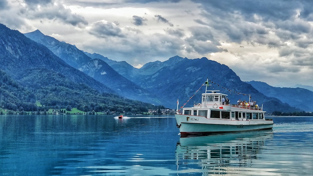 white fishing boat on body of water near mountains under cloudy sky