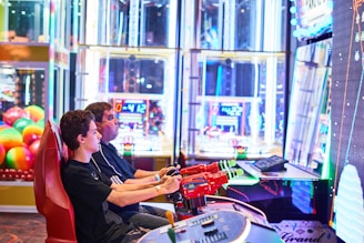 Two people are seated at an arcade game console, each holding onto red plastic guns aimed at a large, brightly lit screen. The background is filled with colorful arcade lights and gaming machines, creating a vivid and playful atmosphere.