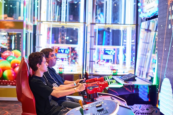 Two people are seated at an arcade game console, each holding onto red plastic guns aimed at a large, brightly lit screen. The background is filled with colorful arcade lights and gaming machines, creating a vivid and playful atmosphere.