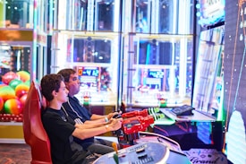 Two people are seated at an arcade game console, each holding onto red plastic guns aimed at a large, brightly lit screen. The background is filled with colorful arcade lights and gaming machines, creating a vivid and playful atmosphere.