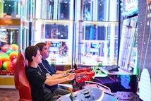 Two people are seated at an arcade game console, each holding onto red plastic guns aimed at a large, brightly lit screen. The background is filled with colorful arcade lights and gaming machines, creating a vivid and playful atmosphere.