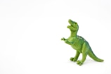 A small, green plastic dinosaur toy is positioned against a plain white background. The figurine resembles a Tyrannosaurus Rex, with its mouth open and arms bent inward.