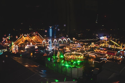 A lively amusement park filled with bright lights illuminates the night. Various attractions, including large tents, roller coasters, and ride structures, can be seen, adorned with colorful light displays. Strings of lights stretch across the park, adding to the festive atmosphere. There are numerous booths and rides scattered throughout the park, casting vibrant reflections.