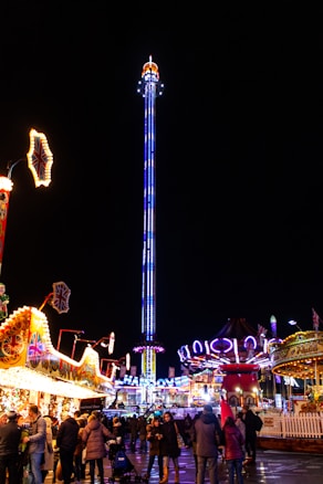 A lively nighttime carnival scene with a tall illuminated tower ride in the center, surrounded by various colorful stalls and attractions. The area is bustling with people, including families and groups enjoying the atmosphere. Bright lights and vibrant colors contribute to the festive environment.