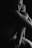 A black and white image of a nude person sitting curled up in a fetal position. Their face is partially visible, with the rest of the body mostly in shadow, creating a dramatic and intimate atmosphere. The lighting highlights the contours of the body, adding depth and emotion.