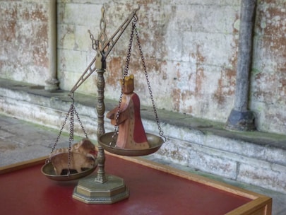 An antique balance scale with a medieval style figure on one side and a carved animal figure resembling a boar or pig on the other. The scale is set against a background of weathered stone walls and placed on a wooden table with a red surface.